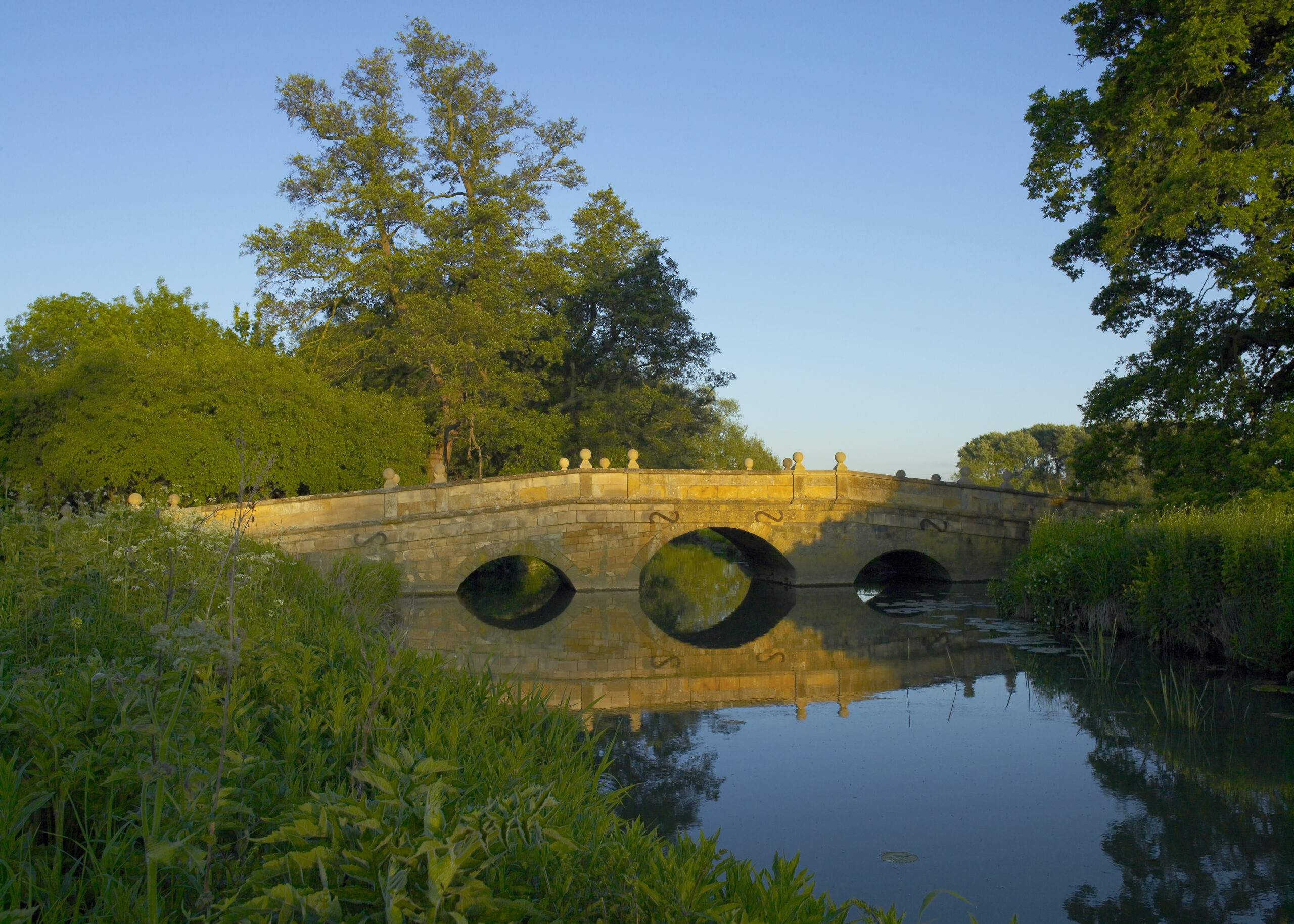 Small bridge made of honey coloured stone with reflection in river Stour